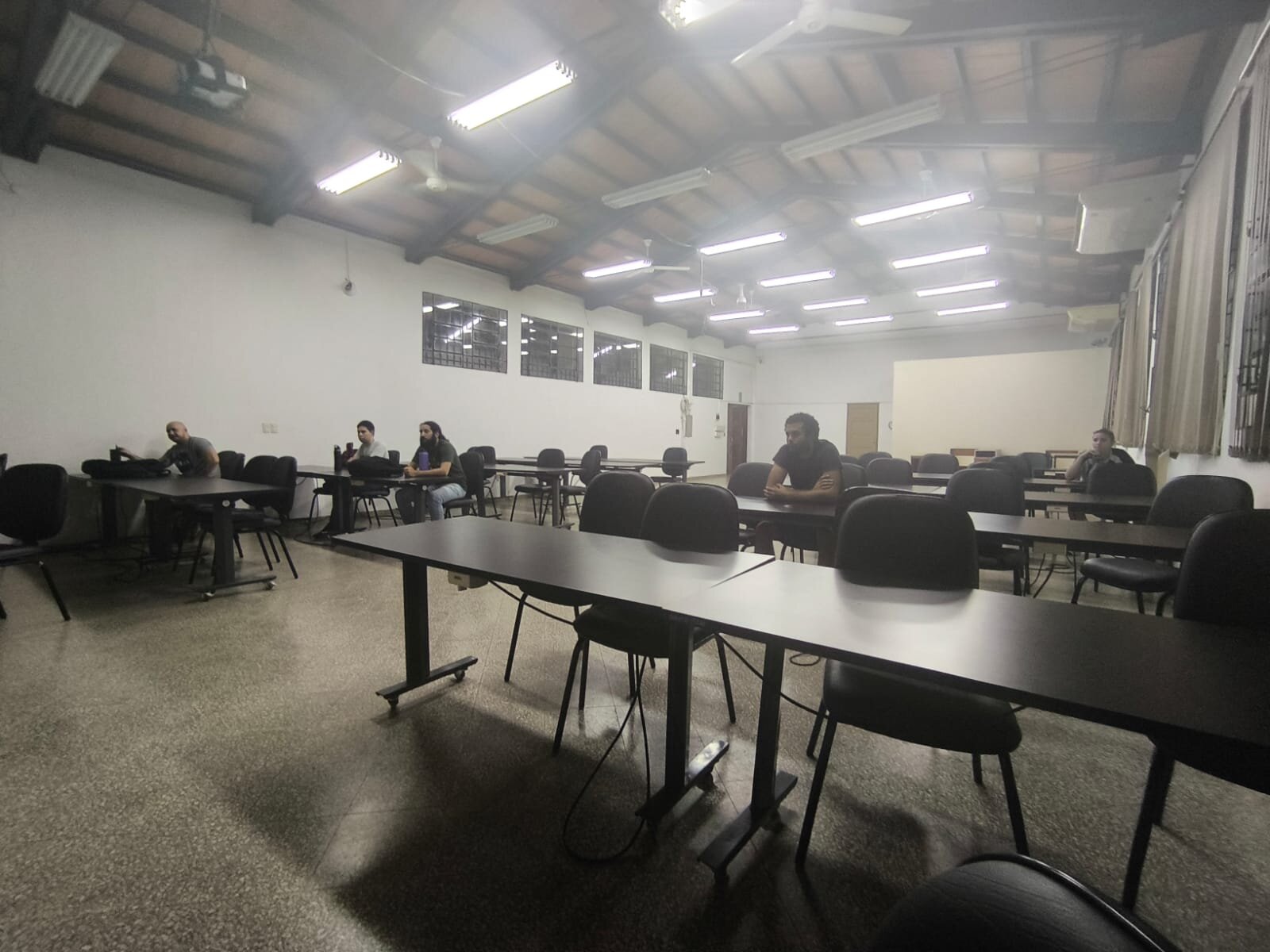 (Photo) A wide-angle photograph of a sparsely populated classroom featuring rows of dark tables and chairs under a slanted ceiling with bright fluorescent lights. people in a classroom | indoor | slanted ceiling, rows of tables, fluorescent lighting | candid | Colors: #7e7e7e, #ffffff, #1a1a1a Note: The image is a real-life photograph capturing an indoor scene with people and furniture.