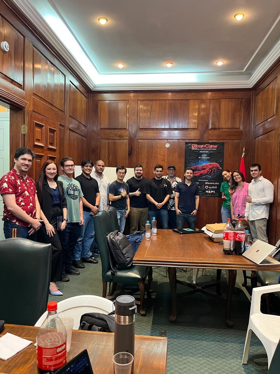 A group of thirteen people pose for a photo in a wood-paneled room around a large conference table, with an event poster in the background.. Text: OpenClaw GLOBAL UNHACKATHON ASUNCIÓN AI Tinkerers SPONSORS ElevenLabs auth0 VAPI Coca-Cola