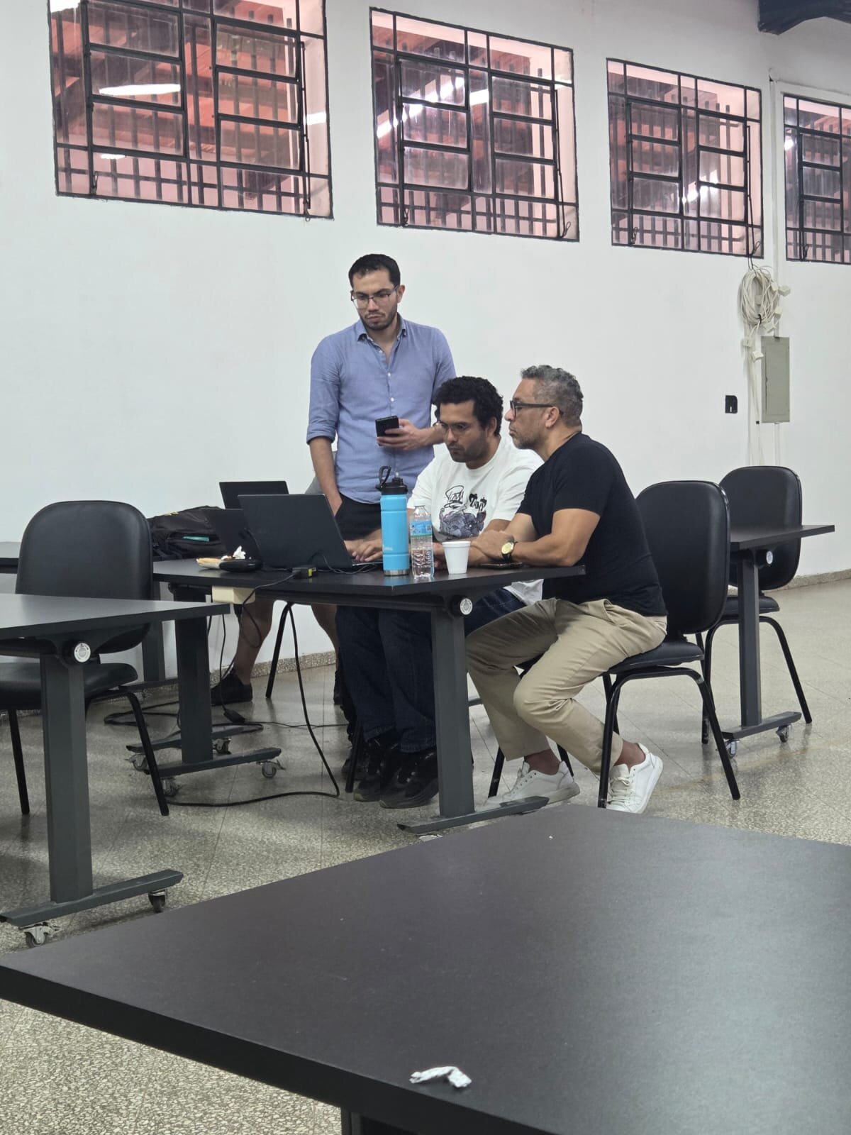 (Photo) Three men are gathered around a desk with laptops in an indoor space, likely a classroom or office setting. people working | indoor | laptops, water bottle, barred windows, tiled floor | candid | Colors: #F5F5F5, #222222, #7A89C2, #D2B48C, #C0C0C0 Note: The image is a real-life photograph capturing a candid moment between people in an indoor environment.