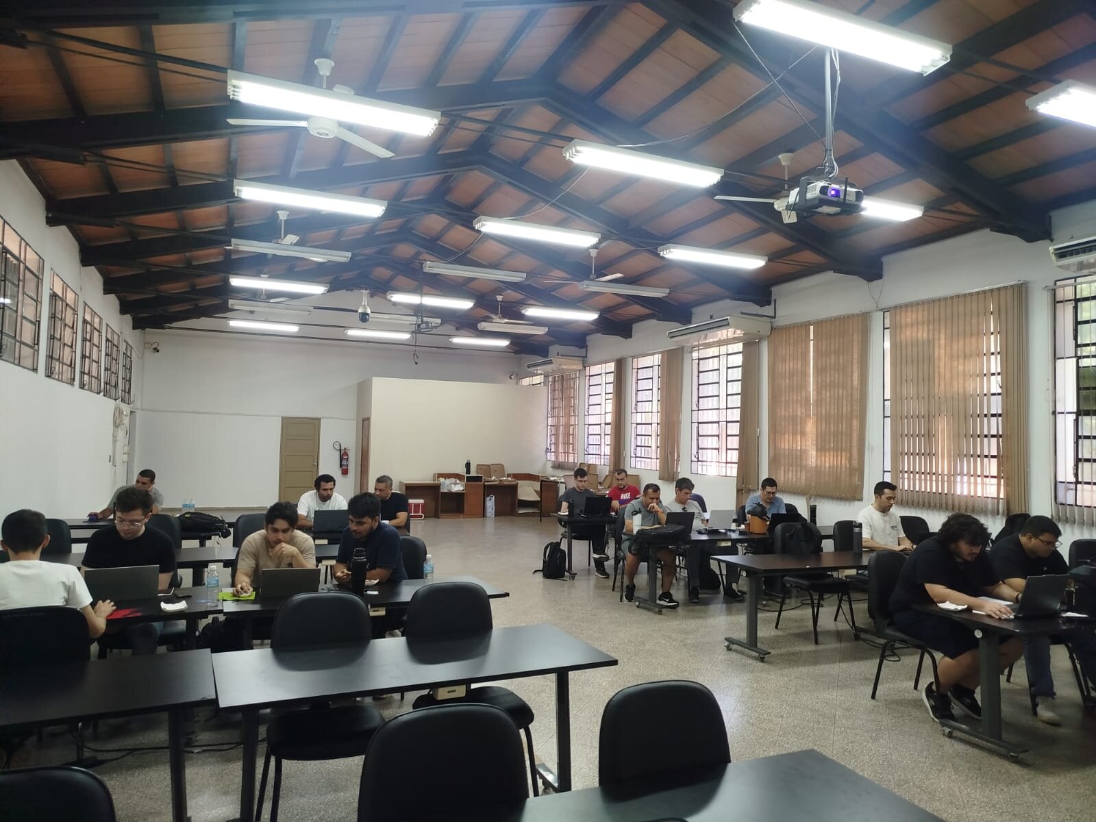 (Photo) A group of people are seated at tables in a large room with a high wooden ceiling, all focused on their laptops in what appears to be a study session or workshop. people working on laptops | indoor room | high wooden ceiling, fluorescent lighting, multiple desks, laptops | candid | Colors: #3B3B3B, #A8A8A8, #6B4E31, #FFFFFF Note: The image is a real-life photograph capturing a group of people in an indoor setting with natural lighting and realistic details.
