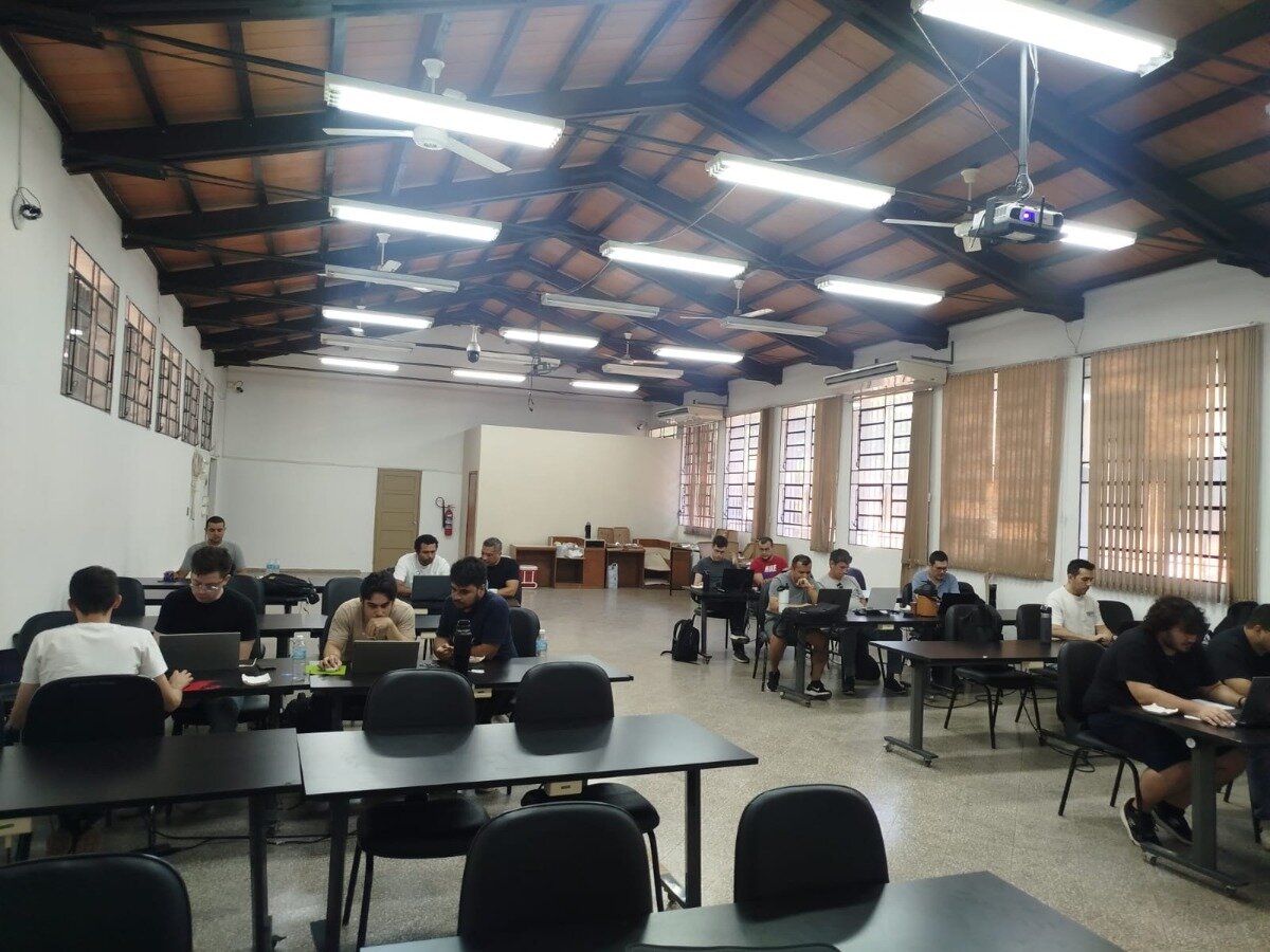 (Photo) A wide-angle photograph of a large, indoor room with a high wooden truss ceiling where several people are seated at tables, most of them working on laptops. people working on laptops | indoor workshop or classroom | high wooden truss ceiling, fluorescent lighting, rows of black tables, projector | candid wide-angle shot | Colors: #3b3b3b, #a0a0a0, #ffffff, #8b4513 Note: The image is a real-life photograph capturing a group of people in an indoor environment.