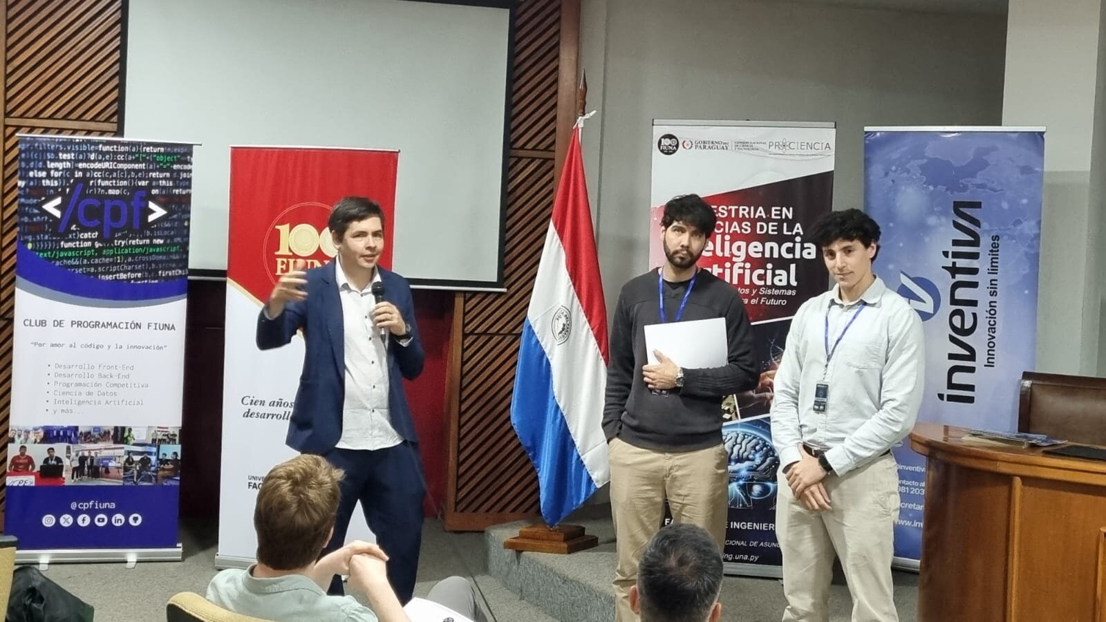 (Photo) A man in a blue suit is giving a presentation at an event, standing in front of banners and a Paraguayan flag, with two other men beside him. Text: CLUB DE PROGRAMACIÓN FIUNA, </cpf>, Por amor al código y la innovación, Desarrollo Front-End, Desarrollo Back-End, Programación Competitiva, Ciencia de Datos, Inteligencia Artificial, y más., @cpfiuna, 100 FIUNA, Cien años desarrollando..., GOBIERNO DEL PARAGUAY, PROCIENCIA, MAESTRÍA EN CIENCIAS DE LA INTELIGENCIA ARTIFICIAL, Modelos y Sistemas para el Futuro, FACULTAD DE INGENIERÍA, UNIVERSIDAD NACIONAL DE ASUNCIÓN, www.ing.una.py, inventiva, Innovación sin límites, www.inventiva.com.py people at a presentation | indoor | multiple banners, Paraguayan flag, speaker with microphone | candid | Colors: #FFFFFF, #0000FF, #FF0000, #2E3192 Note: This is a real-life photograph capturing a moment during a presentation or event with people and physical objects.