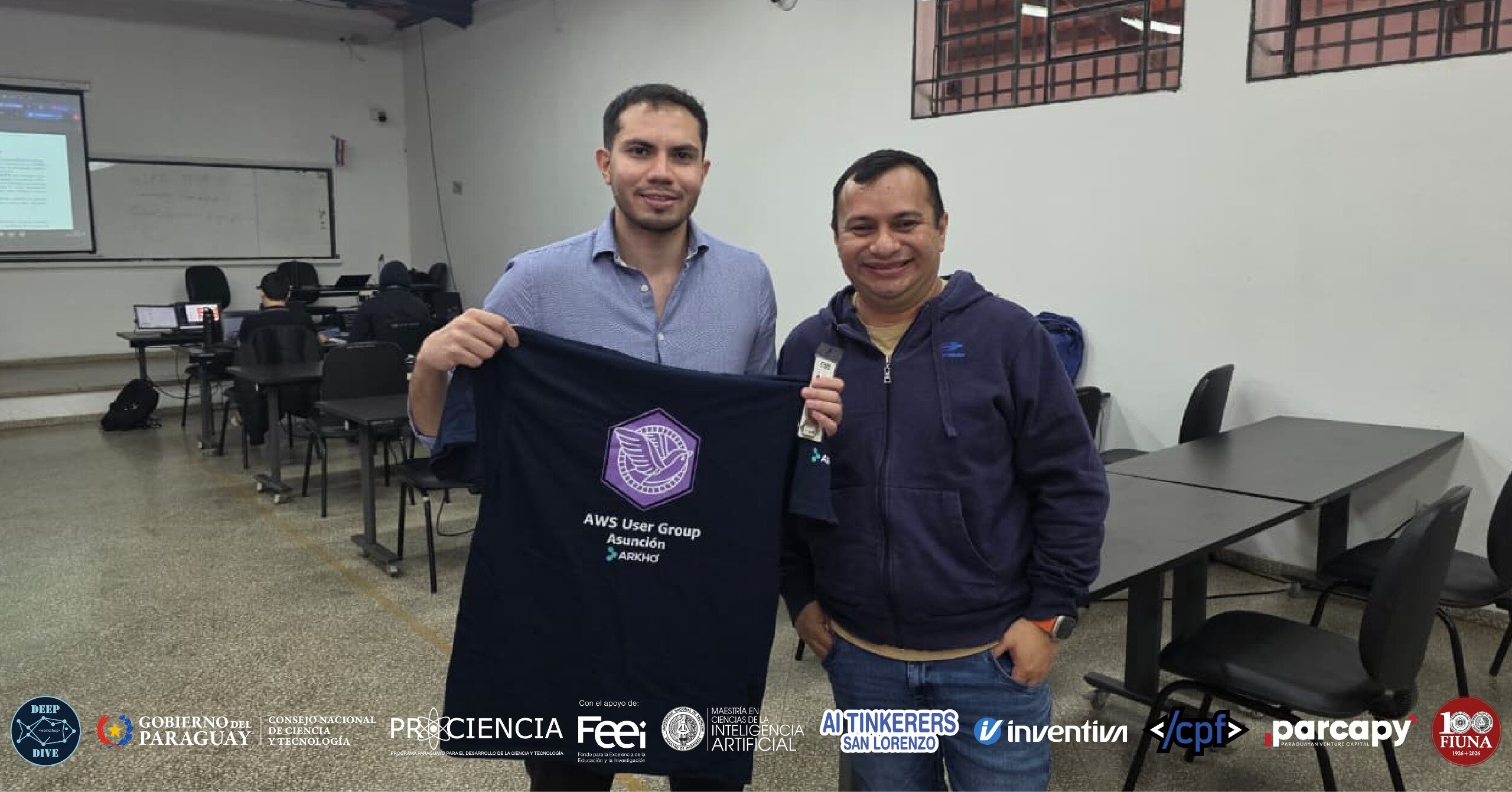 (Photo) Two men pose in a classroom setting, with one holding up a black t-shirt featuring the AWS User Group Asunción logo. The image includes a banner of various organizational logos at the bottom. Text: DEEP DIVE, GOBIERNO DEL PARAGUAY, CONSEJO NACIONAL DE CIENCIA Y TECNOLOGÍA, PRO CIENCIA, Con el apoyo de: Feei, MAESTRÍA EN CIENCIAS DE LA INTELIGENCIA ARTIFICIAL, AI TINKERERS SAN LORENZO, inventiva, </cpf>, parcapy, 100 FIUNA, AWS User Group Asunción, ARKHO people | indoor classroom | promotional t-shirt, organizational logos banner, classroom background with desks and a projector screen | candid portrait | Colors: #f4f4f4, #1c1c1c, #3b4d8c Note: The image is a real-life capture of people in an indoor environment, characteristic of a photograph.