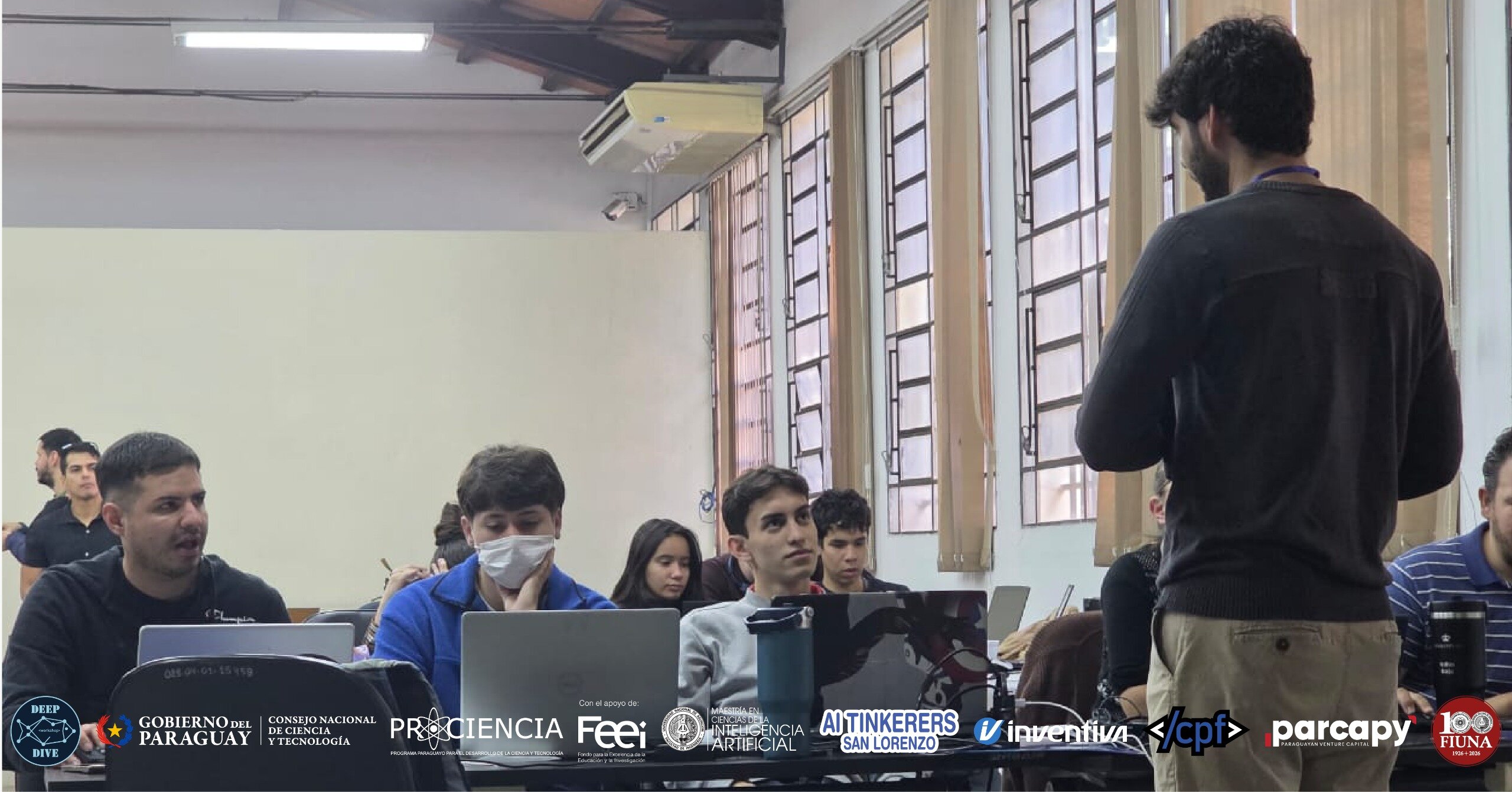 (Photo) A group of people attending a workshop or presentation in an indoor setting, with participants seated at tables with laptops and one person standing in the foreground. Text: DEEP DIVE GOBIERNO DEL PARAGUAY CONSEJO NACIONAL DE CIENCIA Y TECNOLOGÍA PRO CIENCIA Con el apoyo de: Feei MAESTRÍA EN CIENCIAS DE LA COMPUTACIÓN INTELIGENCIA ARTIFICIAL AI TINKERERS SAN LORENZO inventiva </cpf> parcapy 100 FIUNA People in a workshop or classroom | indoor | Multiple people, laptops open, logos at the bottom, one person standing with back to camera | candid | Colors: #FFFFFF, #000000, #D3D3D3, #4A4A4A Note: The image is a real-life photograph capturing a scene of people in an educational or professional environment.