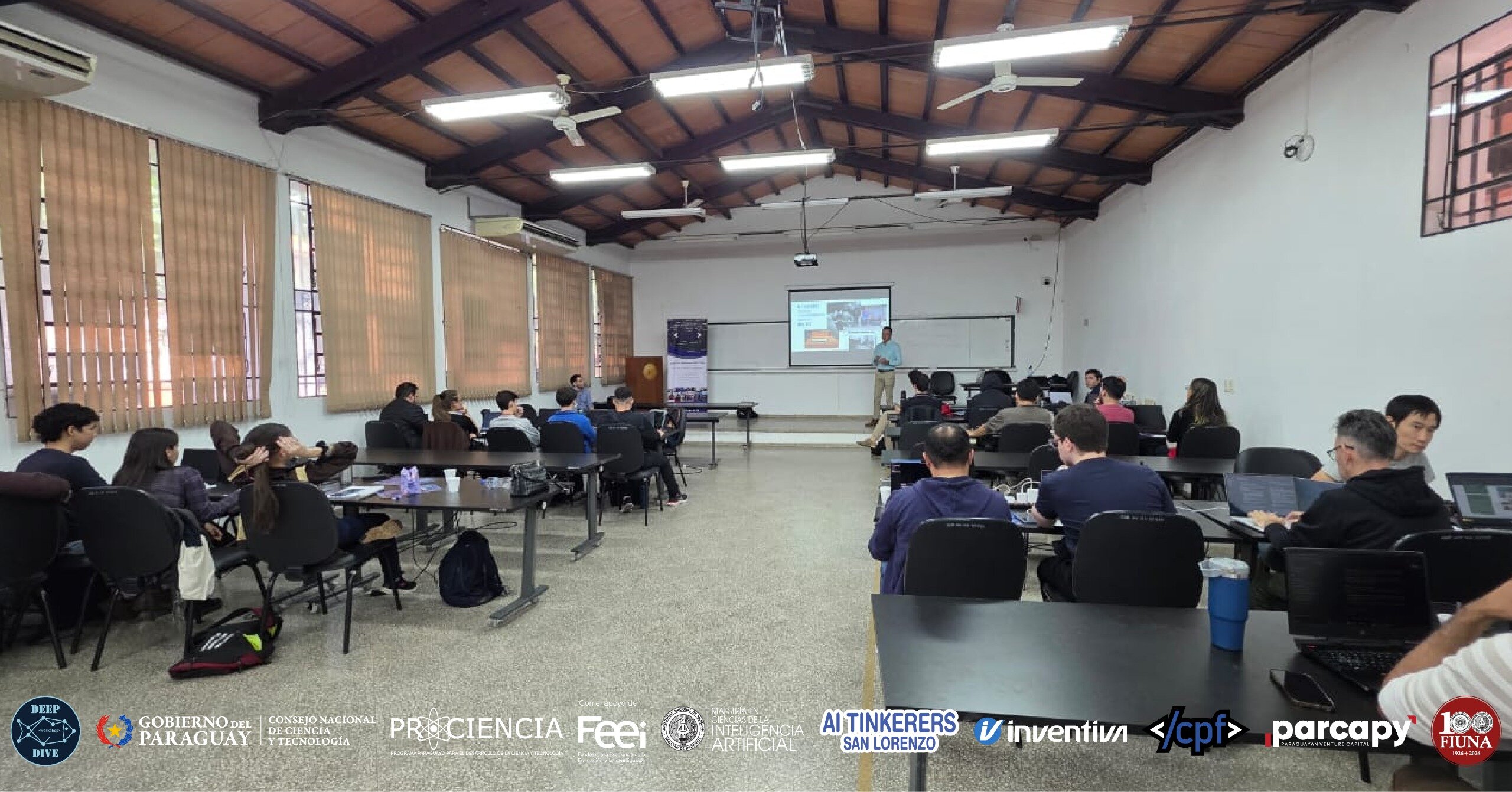 (Photo) A group of people are attending a presentation in a classroom-style setting with a presenter standing at the front near a projector screen. Text: DEEP DIVE GOBIERNO DEL PARAGUAY CONSEJO NACIONAL DE CIENCIA Y TECNOLOGÍA PROCIENCIA FEEI MAESTRÍA EN INTELIGENCIA ARTIFICIAL AI TINKERERS SAN LORENZO inventiva </cpf> parcapy 100 FIUNA presentation | indoor | multiple people, projector screen, laptops, organizational logos at the bottom | candid | Colors: #8B4513, #FFFFFF, #808080, #000000 Note: The image is a photograph capturing a real-life event in an indoor environment, featuring people and physical objects.