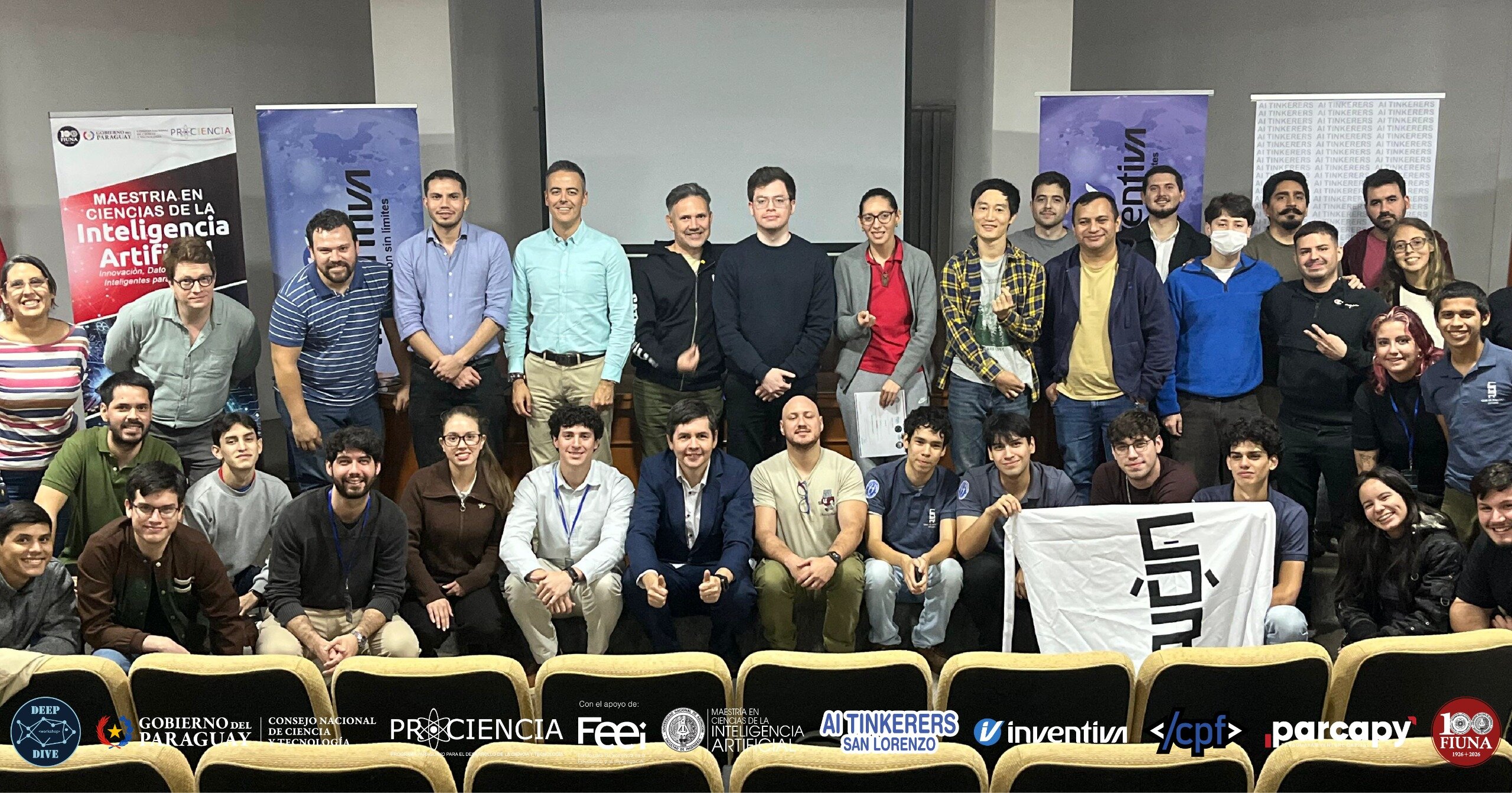A large group of students and faculty posing for a commemorative group photo in an auditorium, featuring several event banners and sponsor logos.. Text: MAESTRÍA EN CIENCIAS DE LA Inteligencia Artificial, Innovación, Datos y Sistemas Inteligentes para..., MVA Innovación sin límites, inventiva, AI TINKERERS, DEEP DIVE, GOBIERNO DEL PARAGUAY, CONSEJO NACIONAL DE CIENCIA Y TECNOLOGÍA, PROCIENCIA, Con el apoyo de: Feei, AI TINKERERS SAN LORENZO, </cpf>, parcapy, 100 FIUNA