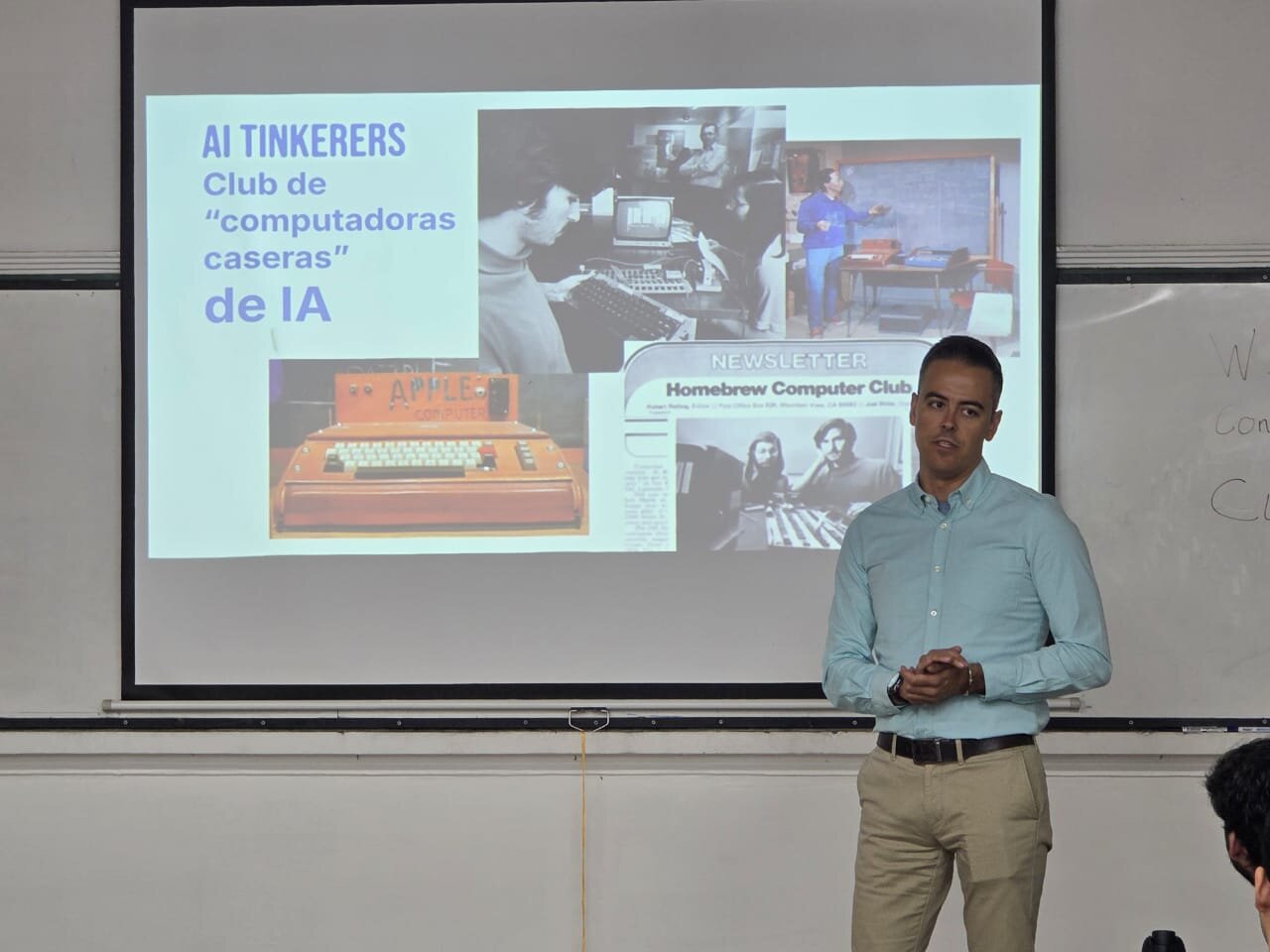 (Photo) A man is standing in front of a projection screen giving a presentation about the history of personal computing and AI tinkerers. Text: AI TINKERERS Club de "computadoras caseras" de IA APPLE COMPUTER NEWSLETTER Homebrew Computer Club [unreadable] person, presentation | indoor, classroom or office | projection screen with historical computer images, speaker in professional-casual attire | candid | Colors: #FFFFFF, #ADD8E6, #C3B091, #000000 Note: The image is a real-world photograph capturing a person delivering a presentation in an indoor environment.