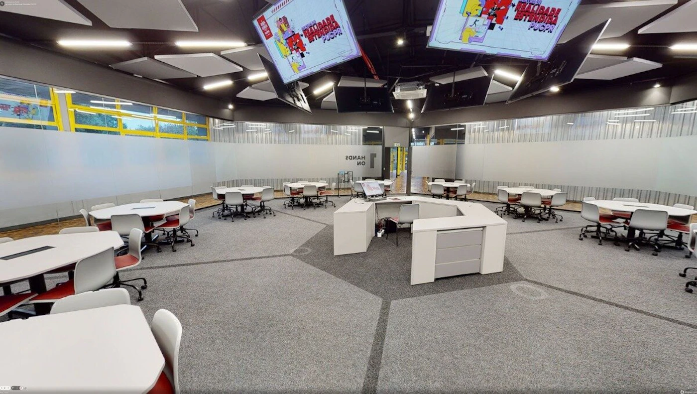 (Photo) A modern, wide-angle photograph of an empty, high-tech classroom or laboratory featuring circular tables, ergonomic chairs, and multiple overhead digital screens. Text: REALIDADE ESTENDIDA PUCPR, HANDS ON Educational or laboratory interior | indoor | Hexagonal floor pattern, overhead monitors, multiple workstations, frosted glass walls | Wide-angle architectural photography | Colors: #D3D3D3, #FFFFFF, #2C2C2C, #B22222 Note: This is a real-world photograph capturing the interior of a physical space with natural lighting and architectural details.